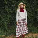 A woman stands outdoors in front of a dense green hedge, wearing a cream knit sweater, a red bow at the collar, a red and green plaid midi skirt, a straw hat, and red socks.