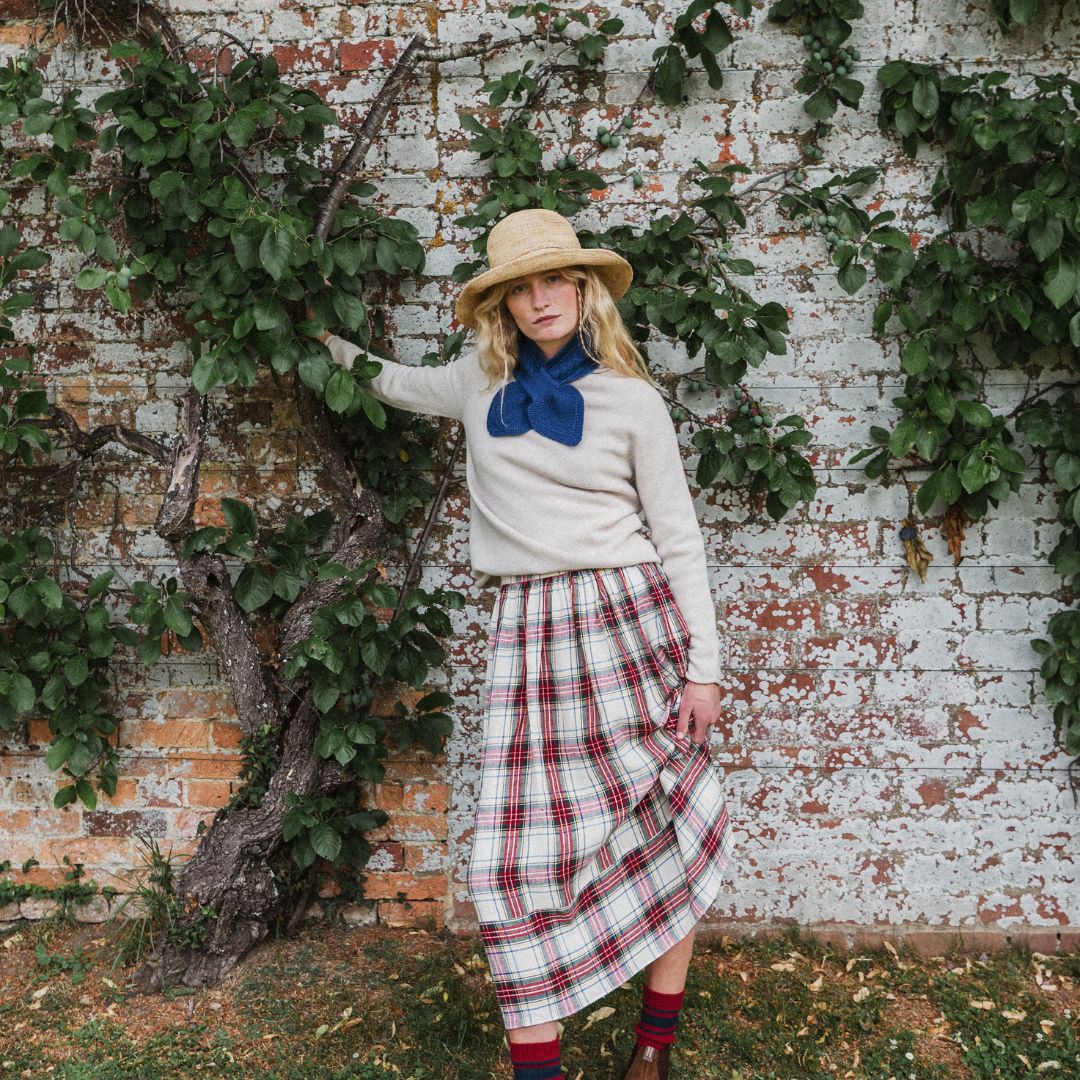 A woman stands outdoors against a weathered brick wall covered with green ivy, wearing a cream sweater, a blue bow tie, a red and white plaid skirt, a straw hat, and red socks.