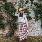 A woman stands outdoors against a weathered brick wall covered with green ivy, wearing a cream sweater, a blue bow tie, a red and white plaid skirt, a straw hat, and red socks.