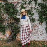 A woman stands outdoors against a weathered brick wall covered with green ivy, wearing a cream sweater, a blue bow tie, a red and white plaid skirt, a straw hat, and red socks.