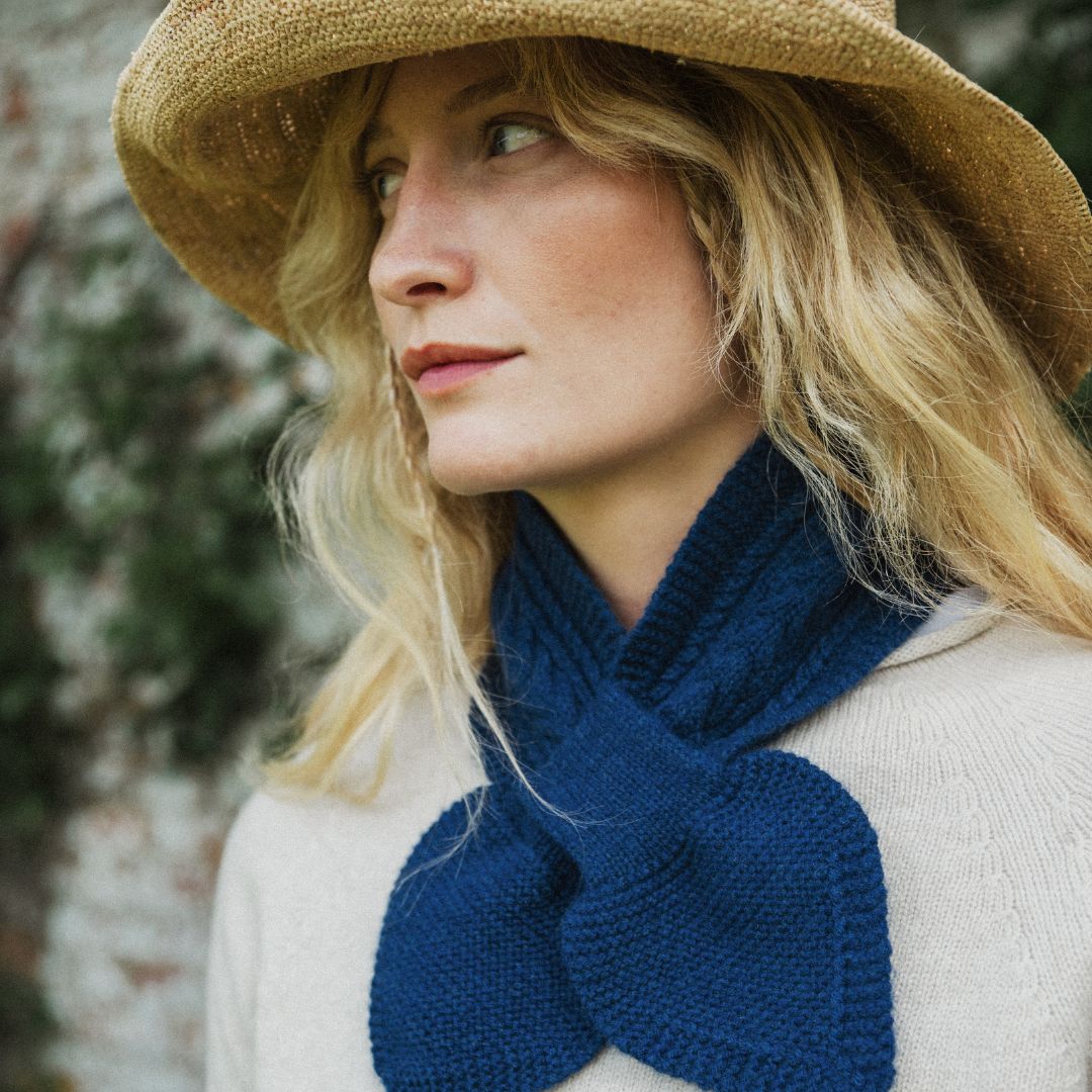 A close-up lifestyle shot of a woman wearing a thick, navy blue knit scarf with a bow detail, a beige straw hat, and a cream-colored knit sweater, set against a soft, blurred outdoor background.