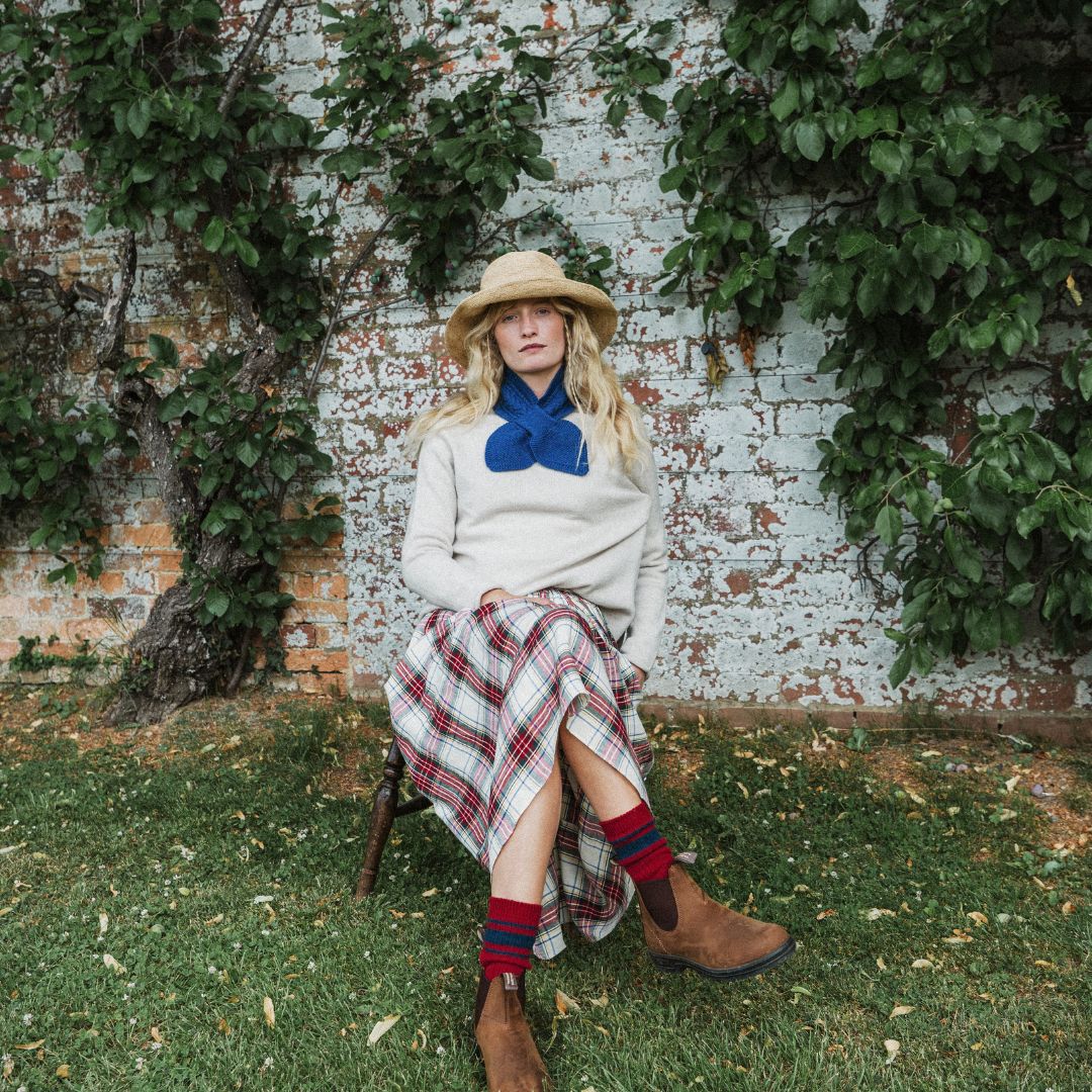 A woman sits on a wooden stool outdoors in front of a brick wall covered in green ivy. She is wearing a cream knit sweater, a large blue bow, a red and white plaid skirt, red and blue striped socks, and brown ankle boots.