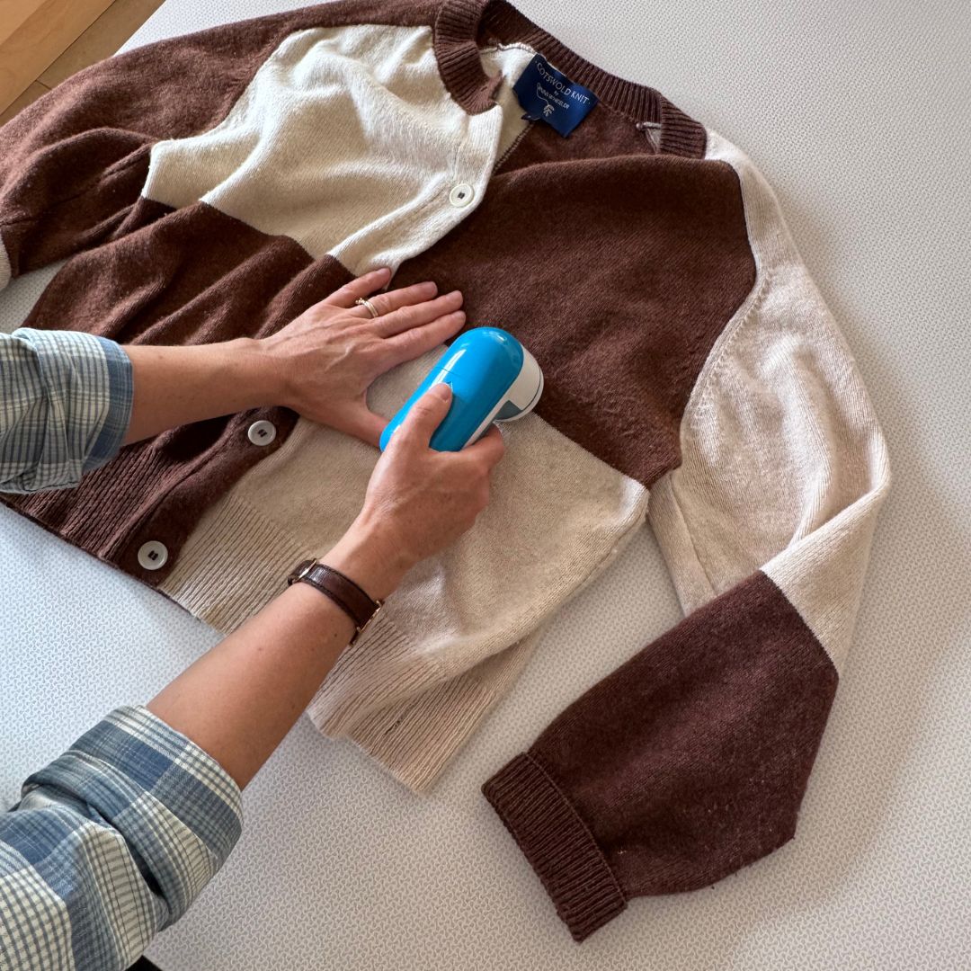 A person uses a blue and white handheld garment pilling maching on a brown and cream colorblock cardigan sweater laid flat on a light-colored textured surface.