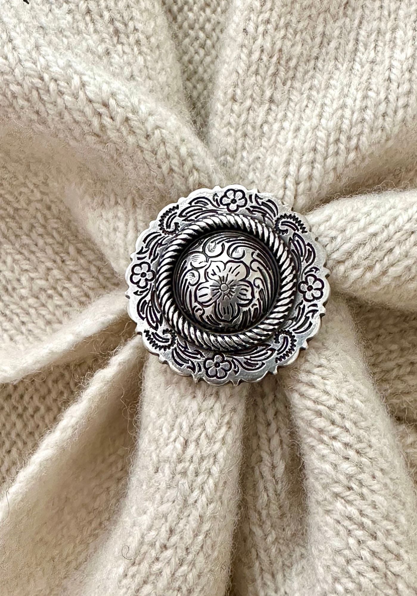 A close-up of a round silver-toned ornate scarf slider with floral and rope-like engraved details, placed over a cream knit fabric background.