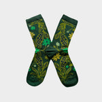 A pair of dark green crew socks with a botanical pattern of yellow and light green leaves and flowers, arranged in an X shape on a light gray background.