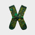 A pair of dark green crew socks with a botanical pattern of yellow and light green leaves and flowers, arranged in an X shape on a light gray background.