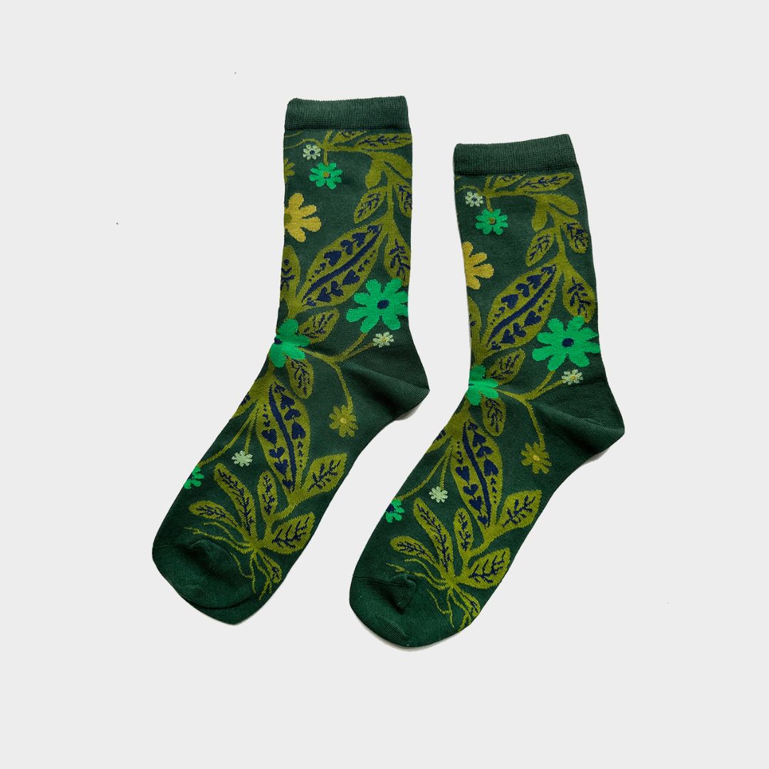 A pair of dark green crew socks with a detailed botanical pattern featuring olive green leaves, lime green flowers, and small yellow accents, displayed against a light gray background.