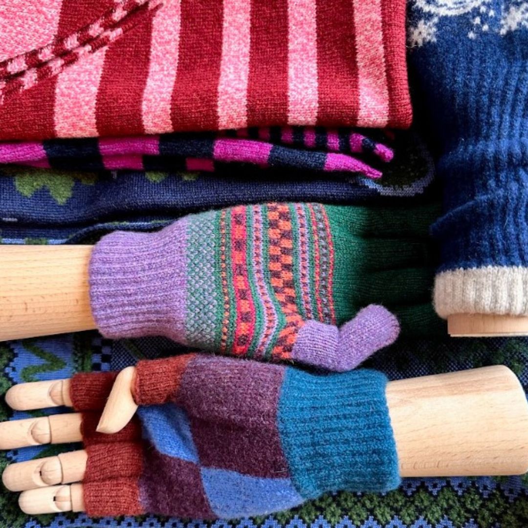 A stack of folded and knitted woolen items displayed on wooden mannequin hands, including striped knitwear in pink and red, a dark blue patterned piece, and two fingerless knit gloves in purple, green, blue, and brown.