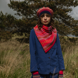Person wearing a blue jacket, red patterned scarf, and colorful hat standing in a grassy field with trees in the background.