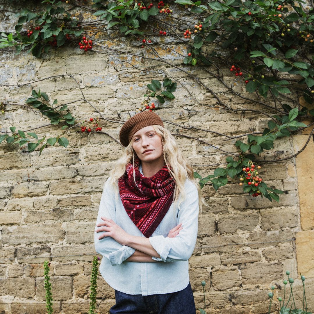 A woman stands with arms crossed in front of a rustic stone wall covered with green ivy and small red berries. She is wearing a brown knit beanie, a light blue button-up shirt, and a dark red patterned scarf.