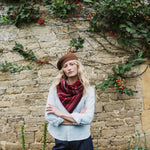 A woman stands with arms crossed in front of a rustic stone wall covered with green ivy and small red berries. She is wearing a brown knit beanie, a light blue button-up shirt, and a dark red patterned scarf.