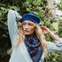 A woman outdoors wearing a bright blue knit beret and a blue patterned knit scarf, with green foliage in the background.