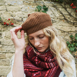 A woman wearing a brown ribbed knit beret and a red and dark pink patterned knit scarf, posing outdoors against a rustic stone wall with small red berries.
