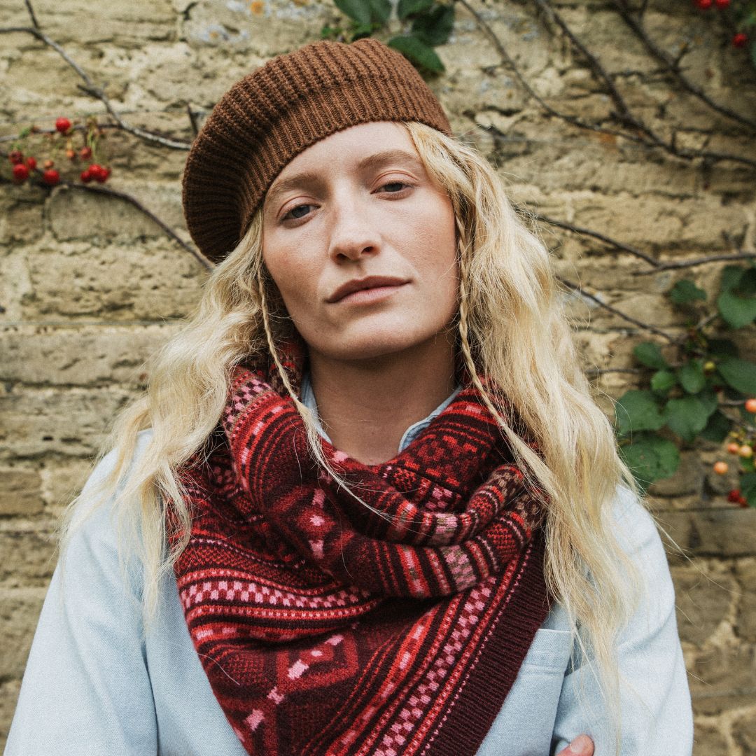 A woman wearing a brown knit beanie and a red and dark brown patterned knit scarf, standing in front of a rustic stone wall with climbing plants.