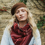 A woman wearing a brown knit beanie and a red and dark brown patterned knit scarf, standing in front of a rustic stone wall with climbing plants.