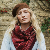 A woman wearing a brown knit beanie and a red and dark brown patterned knit scarf, standing in front of a rustic stone wall with climbing plants.