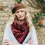 A woman wearing a brown knit beret, a red and dark brown patterned knit scarf, and a light blue cardigan, standing in front of a rustic stone wall with climbing berries.