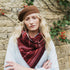 A woman wearing a brown knit beret, a red and dark brown patterned knit scarf, and a light blue cardigan, standing in front of a rustic stone wall with climbing berries.