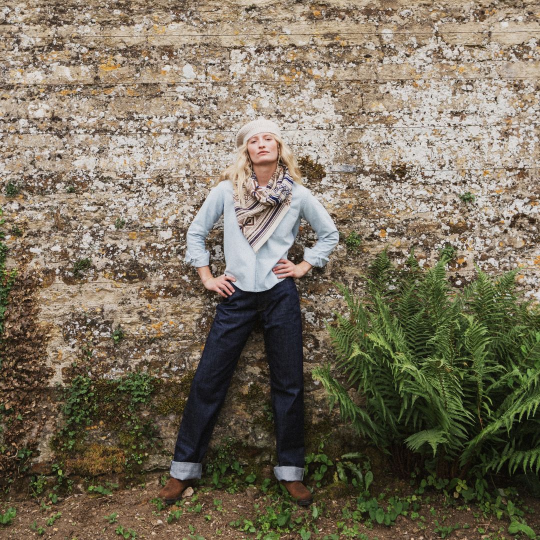 A woman stands against a textured stone wall wearing a light blue long-sleeve shirt, dark blue jeans, a beige and brown patterned scarf, and a cream beret, with green ferns visible on the right.