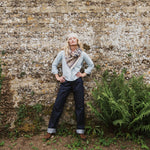 A woman stands against a textured stone wall wearing a light blue long-sleeve shirt, dark blue jeans, a beige and brown patterned scarf, and a cream beret, with green ferns visible on the right.