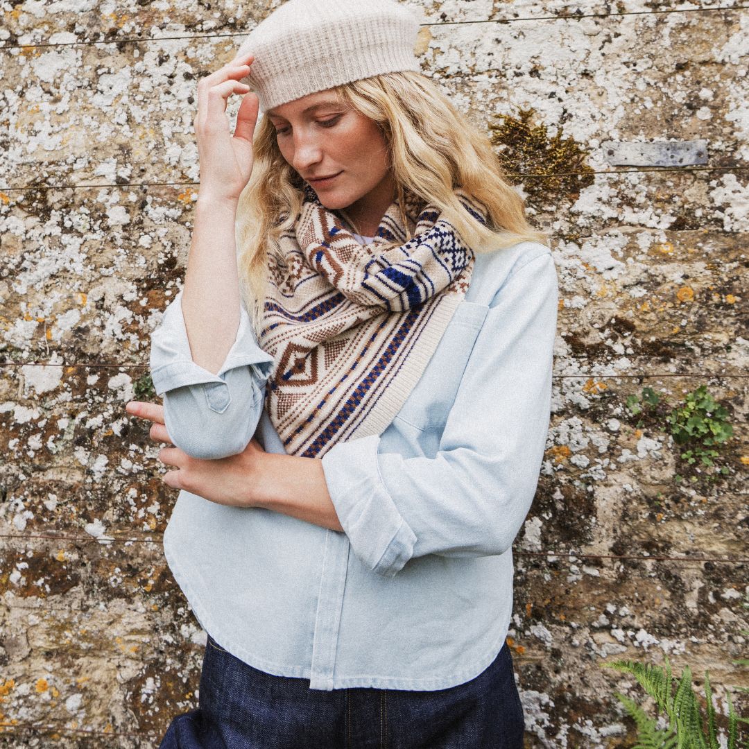 A pwoman wearing a cream knit beret, a patterned knit scarf, a light blue denim shirt, and dark blue jeans, posing against a textured stone wall.
