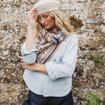A pwoman wearing a cream knit beret, a patterned knit scarf, a light blue denim shirt, and dark blue jeans, posing against a textured stone wall.