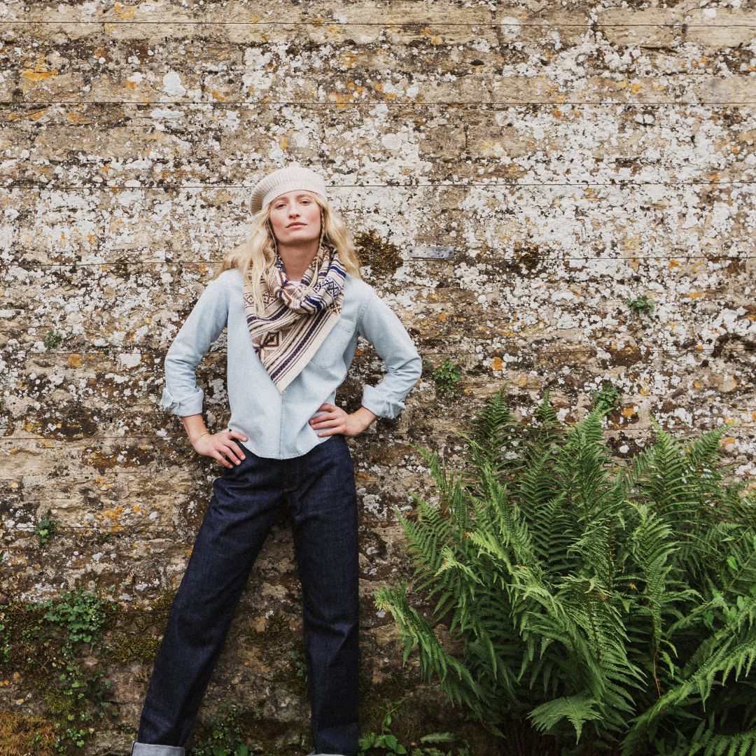 A woman stands against a textured stone wall wearing a light blue shirt, a patterned scarf, dark jeans, and a cream beret, with green ferns in the foreground.