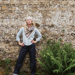 A woman stands against a textured stone wall wearing a light blue shirt, a patterned scarf, dark jeans, and a cream beret, with green ferns in the foreground.