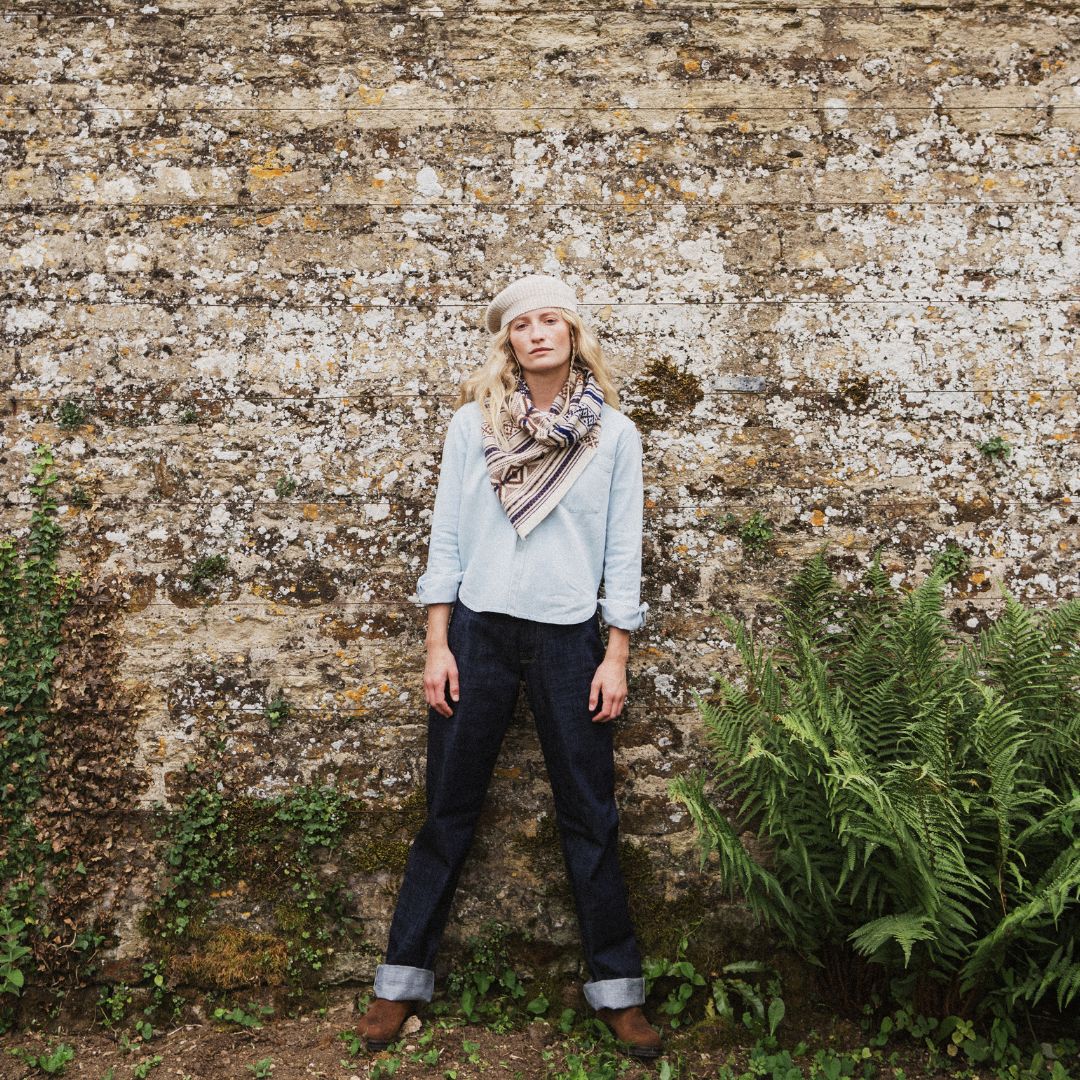A woman stands in front of a weathered stone wall wearing a light blue long-sleeve shirt, dark blue jeans, brown boots, a cream beret, and a striped scarf.