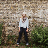 A woman stands in front of a weathered stone wall wearing a light blue long-sleeve shirt, dark blue jeans, brown boots, a cream beret, and a striped scarf.
