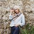 A woman stands against a textured stone wall wearing a light beige knit beret, a light blue denim shirt, a patterned scarf, and dark blue jeans.