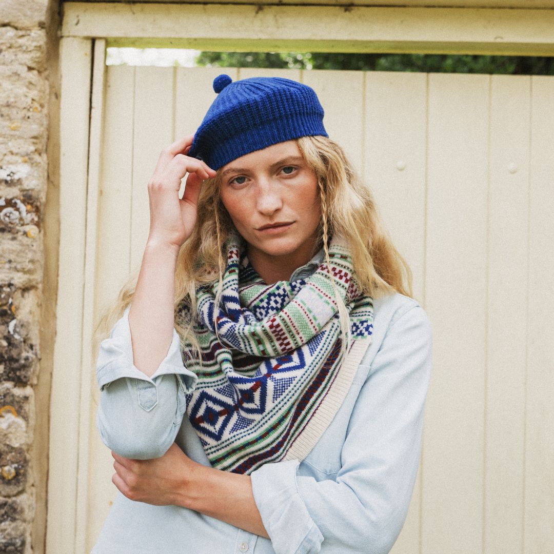 A woman wearing a blue knit beanie with a pom-pom and a multicolor geometric patterned scarf, posing in front of a cream-colored wooden fence.