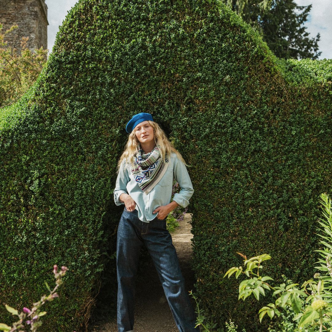 A woman stands in a garden doorway framed by a large trimmed green hedge, wearing a light blue shirt, dark blue jeans, a blue beret, and a patterned scarf.