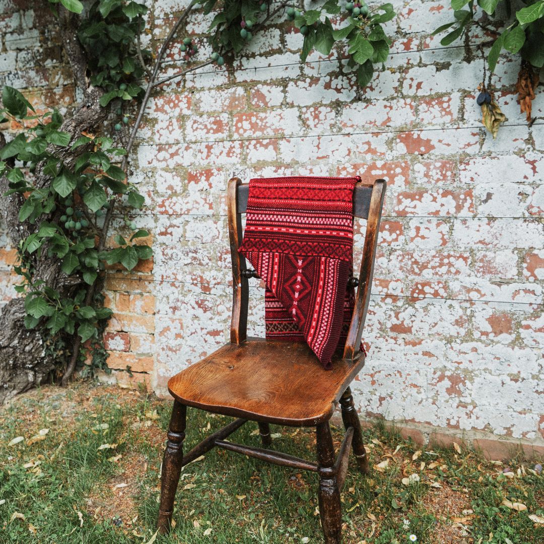 A red and black patterned knit scarf  is draped over a rustic wooden chair placed outdoors in front of a weathered brick wall with climbing green vines.