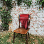 A red and black patterned knit scarf  is draped over a rustic wooden chair placed outdoors in front of a weathered brick wall with climbing green vines.