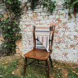 A rustic wooden chair placed outdoors against a weathered brick wall with climbing green vines. A patterned scarf is draped over the chair back and seat.