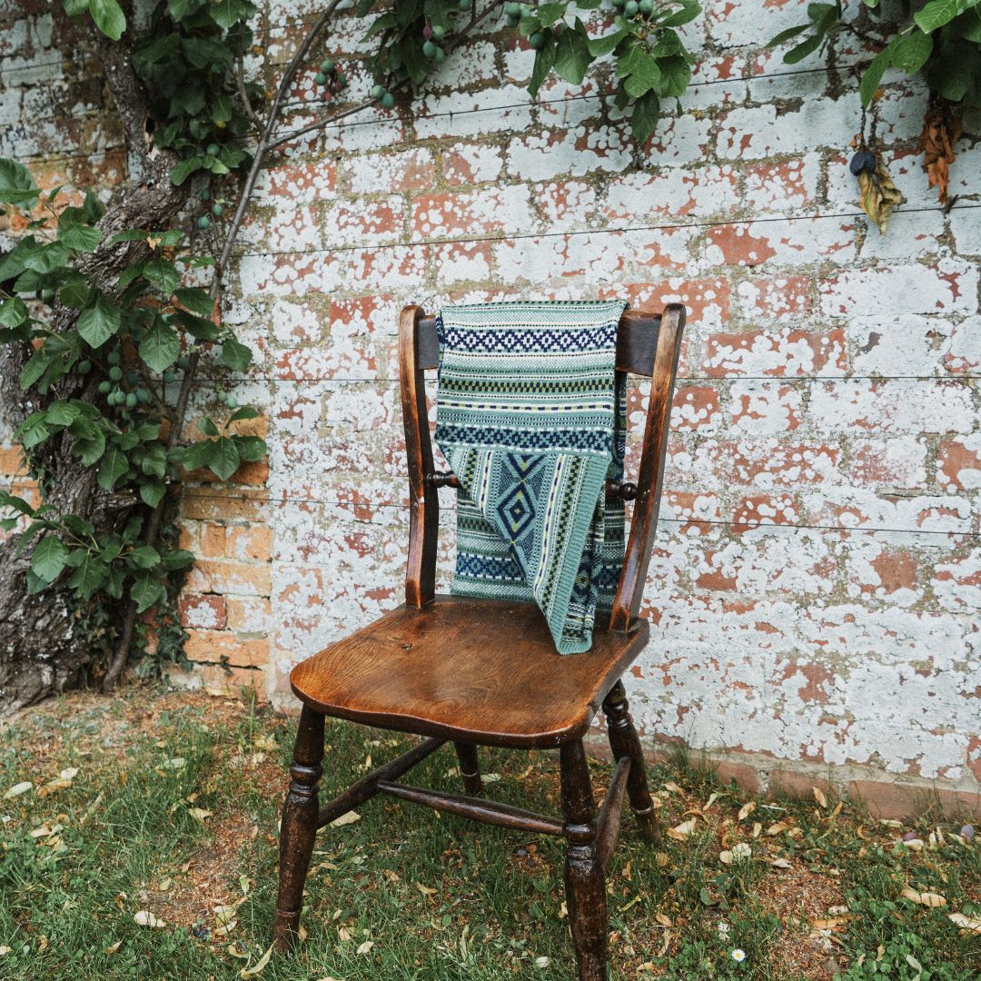 A patterned knit scarf in shades of teal, blue, and cream is draped over a rustic wooden chair set against a weathered brick wall with climbing green vines.