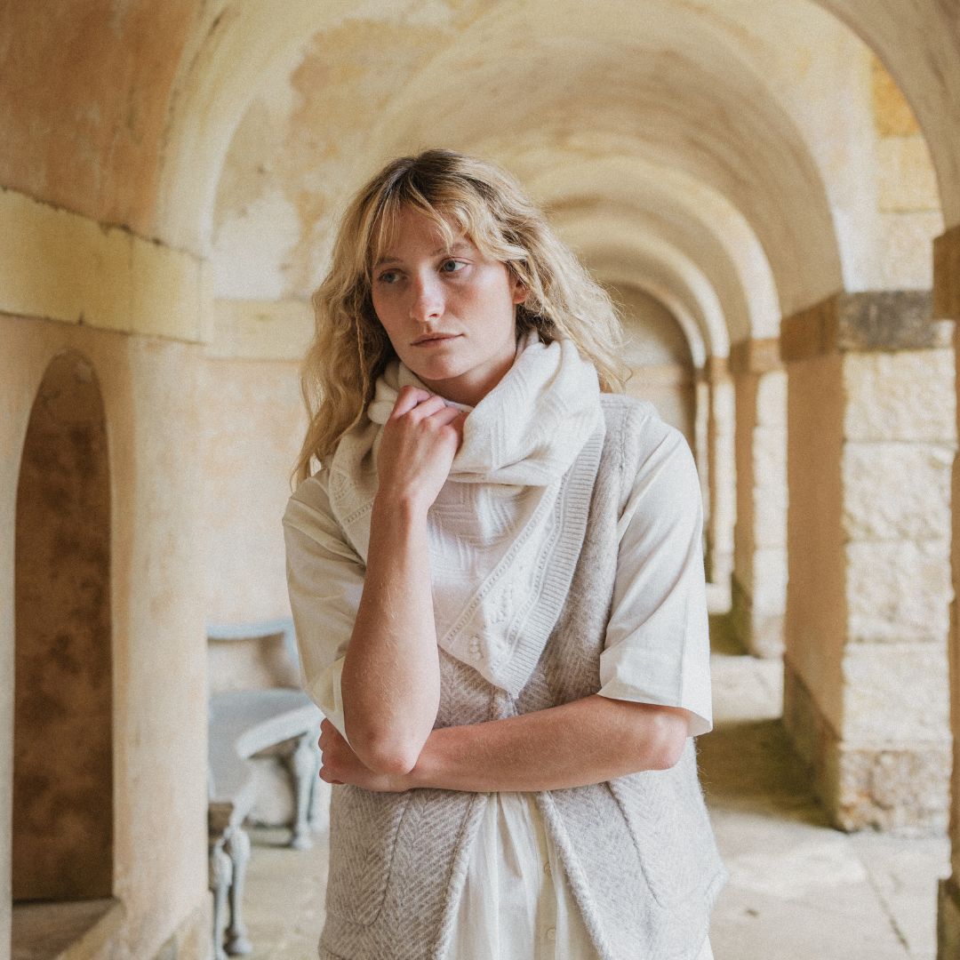 A woman with blonde hair stands in a stone archway corridor wearing a cream-colored textured knit top and matching scarf.