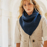 A woman wearing a navy blue knitted triangular scarf or neck warmer over a cream-colored herringbone vest and white shirt, standing in a softly lit architectural setting with arches.