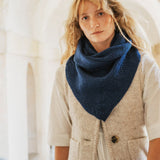 A woman wearing a navy blue knitted triangular scarf or neck warmer over a cream-colored herringbone vest and white shirt, standing in a softly lit architectural setting with arches.