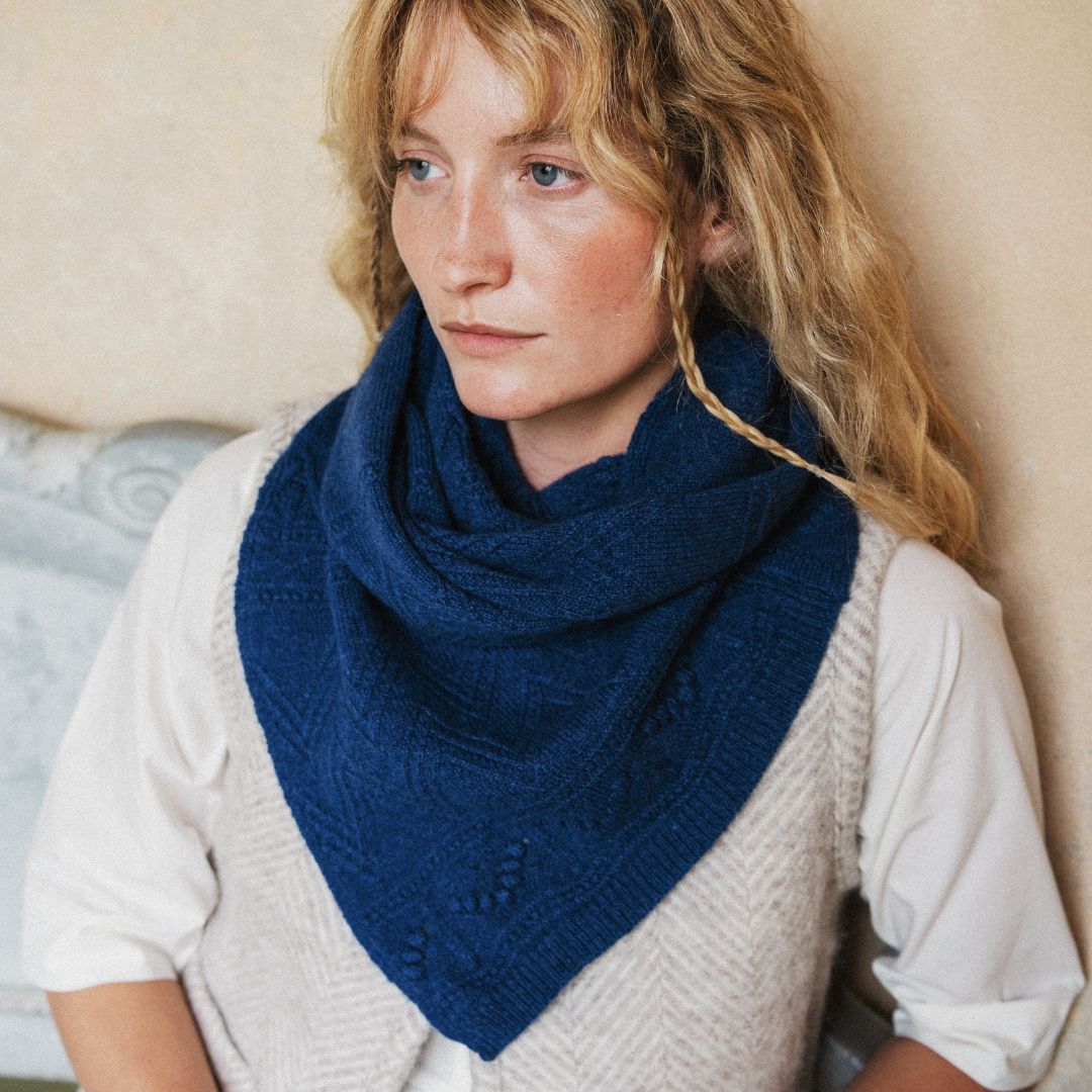 A woman with blonde hair wearing a navy blue knit cowl scarf over a white shirt and light beige vest, leaning against a beige wall.