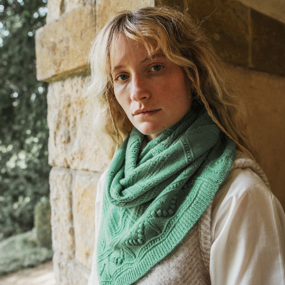 A woman with blonde hair wearing a sage green knitted infinity scarf and a cream-colored sweater vest, leaning against a rustic stone wall.