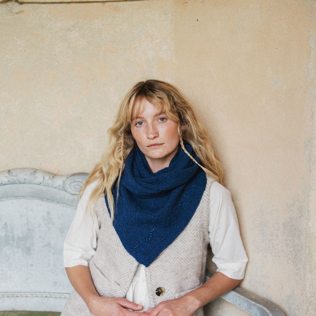 A woman with long blonde hair sits against a textured beige wall, wearing a dark navy blue knit cowl scarf over a white shirt and a light beige herringbone vest.
