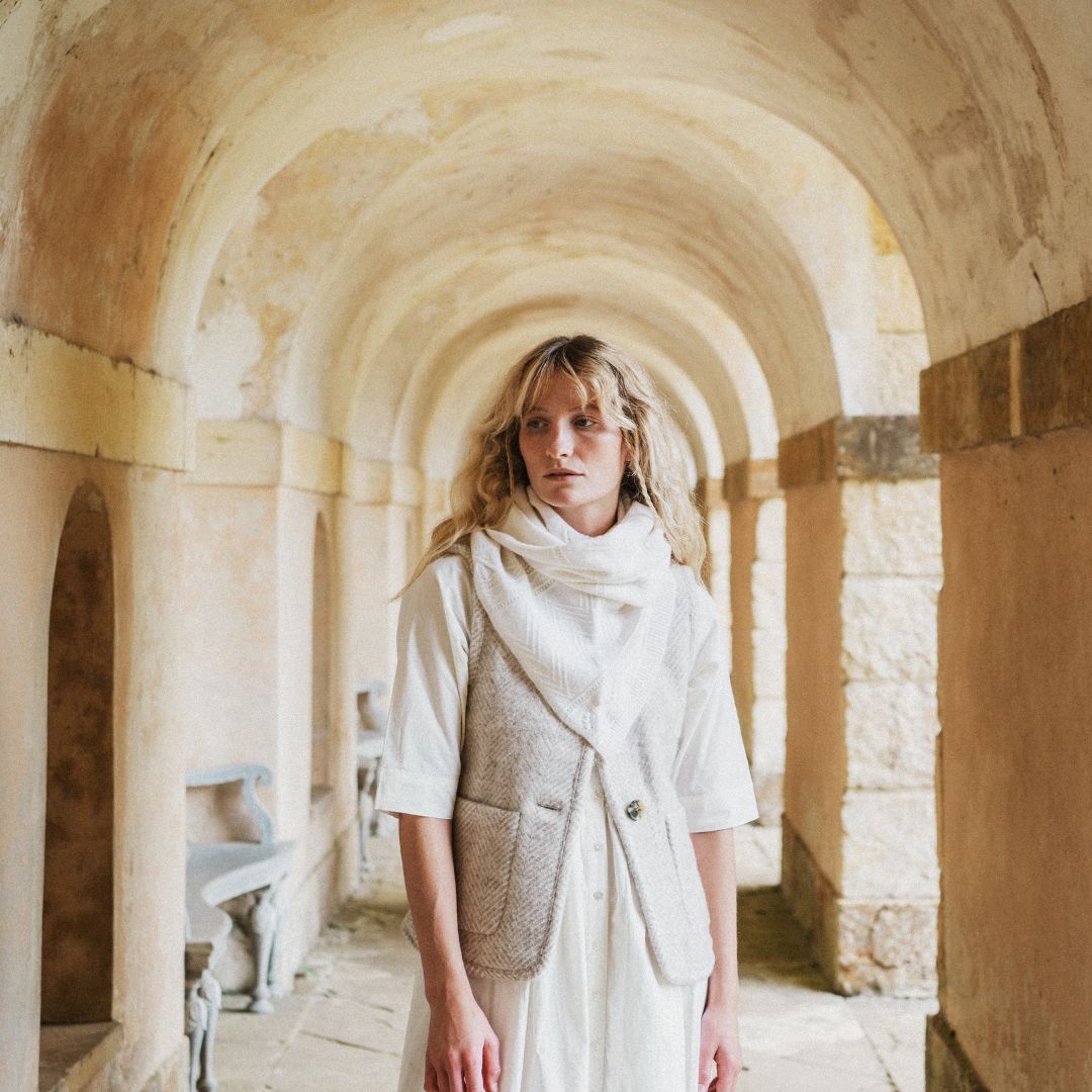 A woman stands in a stone arcade with arched walls, wearing a cream dress, a light scarf, and a textured beige vest.