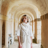 A woman stands in a stone arcade with arched walls, wearing a cream dress, a light scarf, and a textured beige vest.