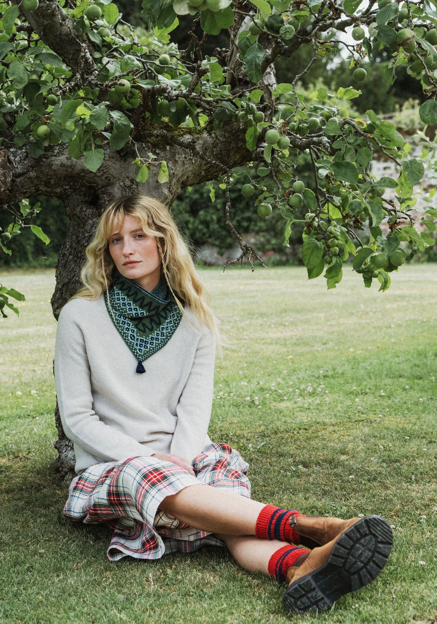 A woman sits on the grass beneath a tree in a park, wearing a cream knit sweater, a green and black patterned twsited snood scarf, a red and white plaid skirt, red socks, and brown boots.