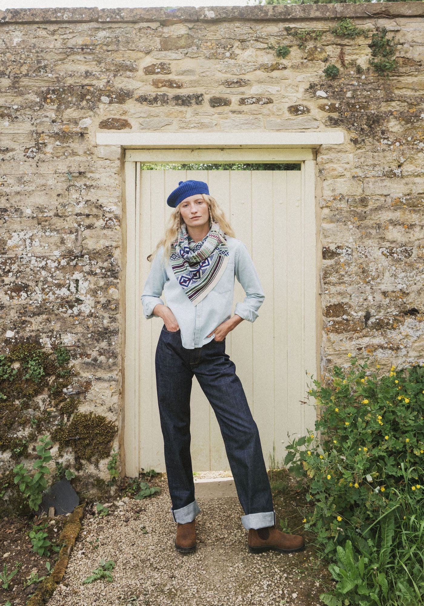 A woman stands in front of a rustic stone wall and cream-colored wooden door, wearing a light blue denim shirt, dark blue jeans, a blue beret, and a patterned tsited snood scarf.
