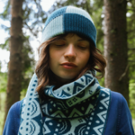 Person wearing a blue and white patterned knit hat and scarf in a forest setting